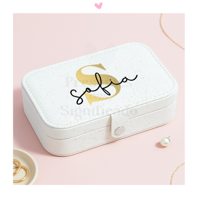 Estojo branco com letra dourada e nome sobre superfície rosa com joias