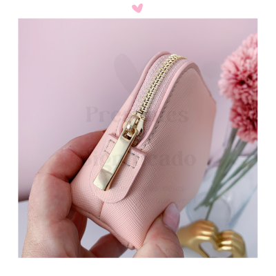 Bolsa pequena rosa com fecho dourado a ser segurada por mãos, com flores e objeto dourado no fundo