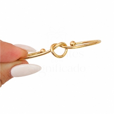 Pulseira dourada com nó e bolas nas extremidades, segurada por dedos com unhas brancas