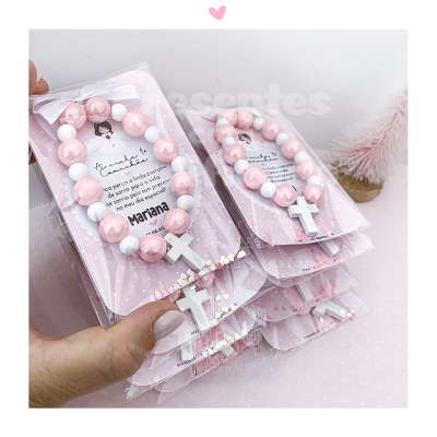 Pulseiras rosa e branco com cruz branca em cartão rosa dentro de embalagem transparente