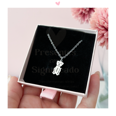 Colar prateado com pendente de urso em caixa preta e branca, mãos seguranado e flores cor-de-rosa ao fundo