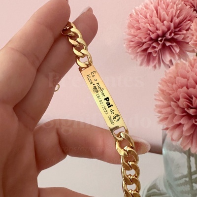 Pulseira dourada com placa gravada e flores cor-de-rosa ao fundo