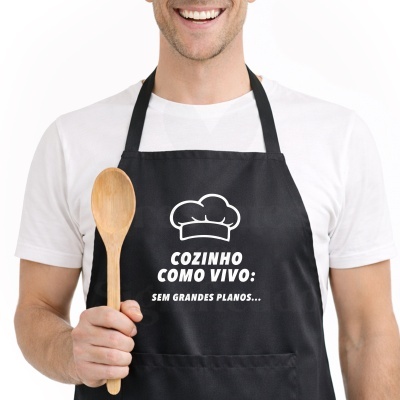 Pessoa com avental preto escrito COZINHO COMO VIVO: SEM GRANDES PLANOS... e segurando uma colher de pau