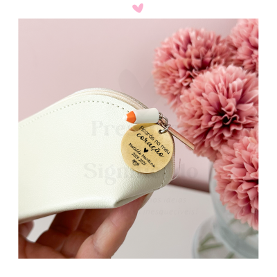 Estojo branco com chaveiro de madeira redondo e lápis laranja, flores cor-de-rosa ao fundo