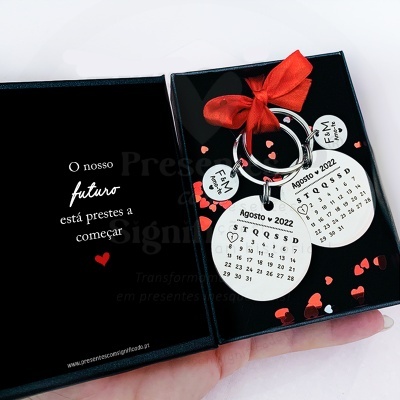 Chaveiros prateados com calendário e pendentes com texto, numa caixa preta com corações e fita vermelha