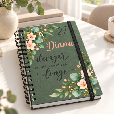 Caderno verde com flores e texto motivacional em português, com fecho elástico preto e espiral, sobre mesa branca.