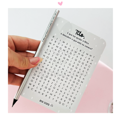 Bloco de palavras cruzadas cinza claro com texto em português sendo segurado por mão, caneta prateada e fundo rosa claro