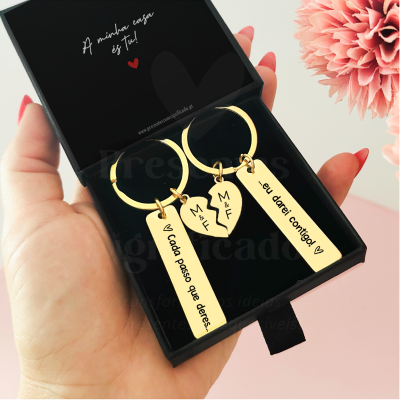 Conjunto de dois porta-chaves dourados com inscrições, em caixa preta, com flor cor-de-rosa ao lado.