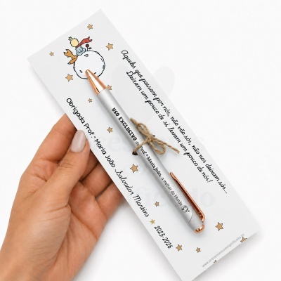 Caneta prateada com detalhes em cobre amarrada a cartolina com ilustração do Pequeno Príncipe e texto em português.