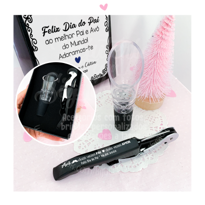 Conjunto de acessórios de vinho com saca-rolhas preto e tampa transparente em caixa preta, com decoração rosa e toalha branca.