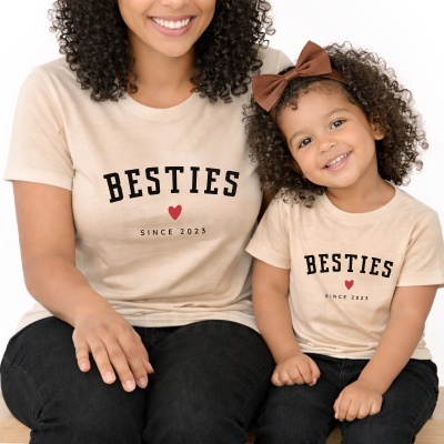 Duas t-shirts bege com texto BESTIES e SINCE 2023 usadas por mulher e criança sentadas.