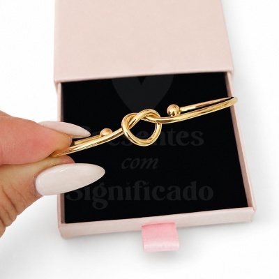 Pulseira de metal dourado com nó central numa caixa de veludo preta e tampa rosa.