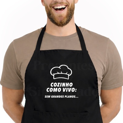 Avental preto com texto em branco COZINHO COMO VIVO: SEM GRANDES PLANOS... usado por pessoa com t-shirt castanha