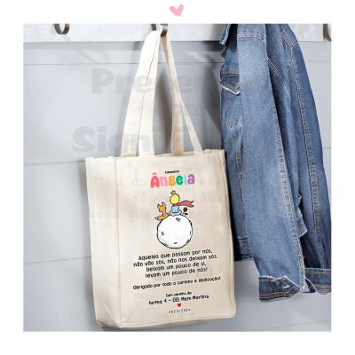 Saco de tecido bege com texto colorido e preto pendurado numa parede cinzenta ao lado de um casaco de ganga