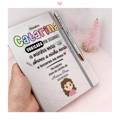 Caderno personalizado cinzento com texto em várias cores e caneta prateada, árvore de Natal rosa ao lado