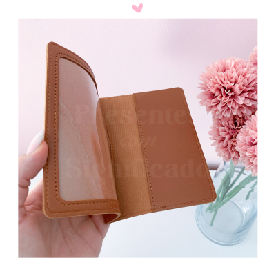 Carteira castanha de couro com compartimento transparente sendo segurada perto de flores cor-de-rosa em vaso