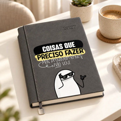Agenda cinzenta com texto humorístico em português numa mesa com planta e café