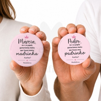 Badges cor de rosa com texto a pedir para serem madrinha e padrinho, seguradas por duas mãos.
