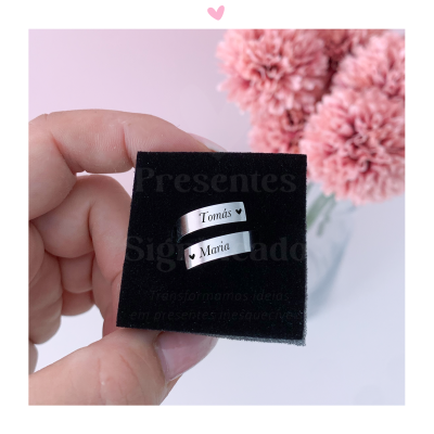 Dois anéis prateados com nomes gravados num suporte de veludo preto com flores ao fundo