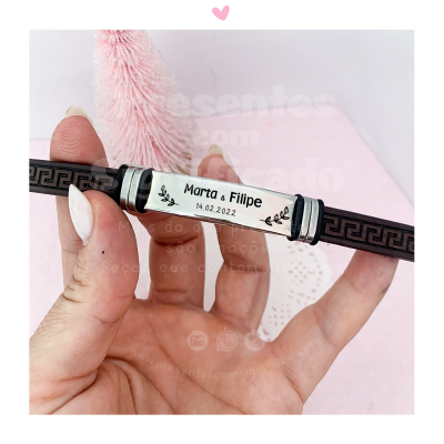 Pulseira preta com placa metálica personalizada contendo nomes e data, segurada por mão com unhas brancas
