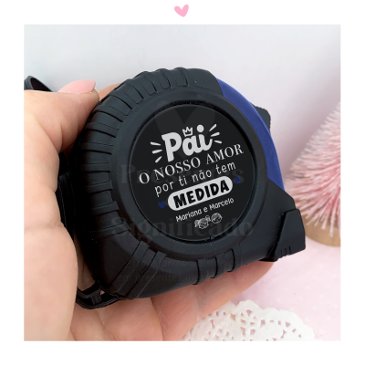 Fita métrica preta com detalhes azuis e texto personalizado sobre amor no centro, segurada por três mãos em ambiente decorado.