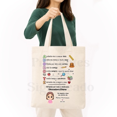 Saco de tecido bege com texto colorido e desenhos escolares, sendo um presente personalizado para educadora.