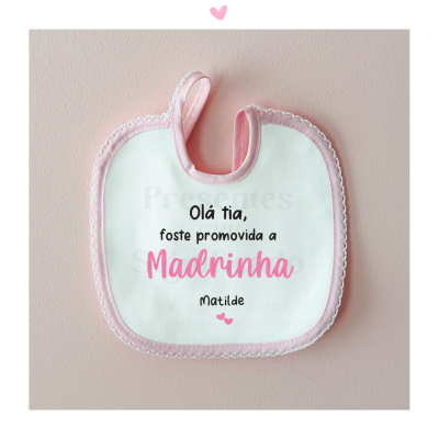 Babete branco com borda rosa e texto declarando promoção a madrinha