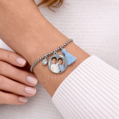 Pulseira prateada com medalhão com foto de duas crianças, cristal e tassel azul claro num pulso com camisola branca de malha.