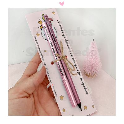 Conjunto de duas canetas rosa em embalagem decorada com estrela e texto, mão segurando, pinheiro rosa ao fundo