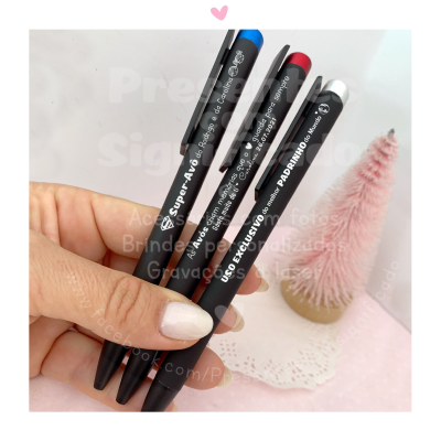 Três canetas pretas com clipes coloridos e texto branco seguradas por mão com unhas brancas, fundo decorativo rosa e árvore rosa