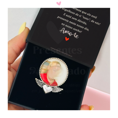 Broche prateado com foto dentro de caixa preta com texto na tampa.