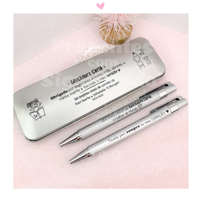 Estojo metálico com duas canetas prateadas e texto impresso em português sobre uma superfície rosa com flor decorativa