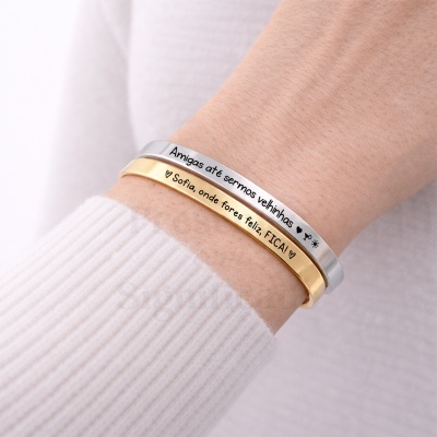 Dois braceletes metálicos, prateado e dourado, com inscrições em português, num pulso com camisola branca.