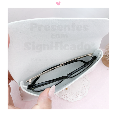 Estojo branco aberto contendo óculos de sol de armação preta e hastes douradas sobre superfície rosa e guardanapo branco.