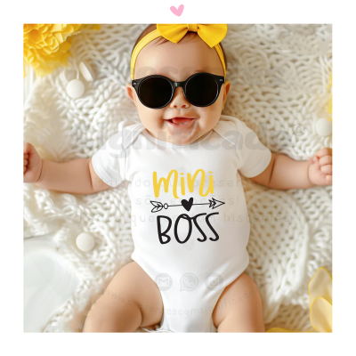 Bebé com body branco 'Mini BOSS' com texto amarelo e preto, óculos de sol e laço amarelo, deitada em manta branca de malha