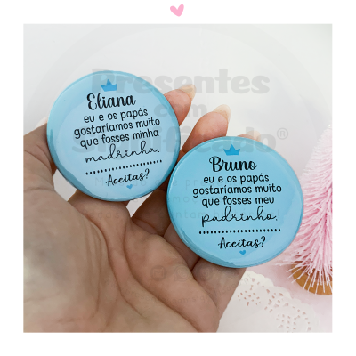 Duas caixas redondas azuis com texto em português sobre pedido para madrinha e padrinho, numa mão, mesa branca e pinheiro rosa