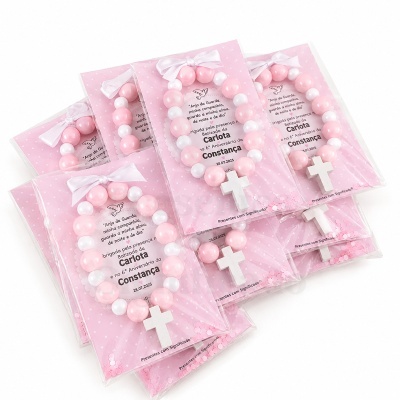 Pulseiras com contas rosas e brancas e cruz branca embaladas em plástico com carta de fundo rosa