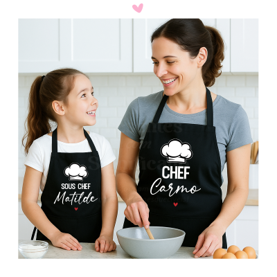 Duas aventais pretas personalizadas com textos e chapéu de chef usadas por mulher e menina numa cozinha branca.