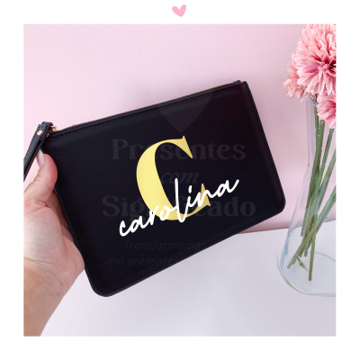 Bolsa preta com a letra C amarela e nome carolina em branco, mão segurando, fundo rosa e vaso de flores