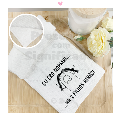 Toalha branca de algodão com texto humorístico e ilustração, sobre superfície de madeira clara com colher de pau, jarro e flores brancas.