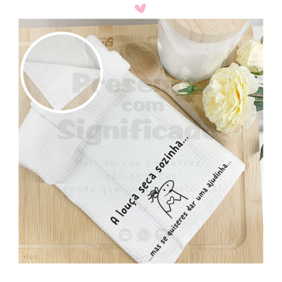 Toalha branca de cozinha com texto bordado e ilustração, sobre superfície de bambu com utensílios e flor.