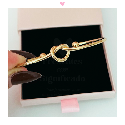 Pulseira dourada com nó e esferas numa caixa de presente rosa.