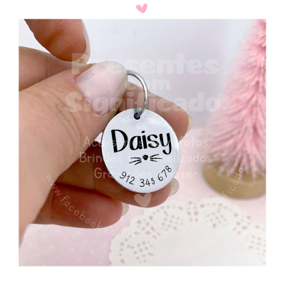 Pingente redondo em metal com nome e número de telefone, contra fundo claro com decoração rosa