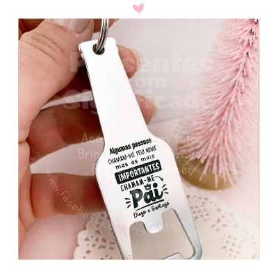 Chaveiro abridor de garrafa metálico prateado com texto em português sobre fundo branco e pinheiro rosa