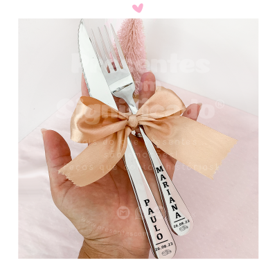 Conjunto de faca e garfo em aço inox com nomes gravados e laço bege segurados por mão