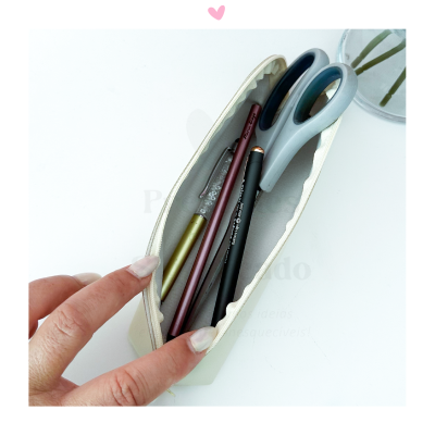 Estojo aberto com tesoura, lápis e canetas, mão segurando, superfície branca e vaso transparente ao fundo