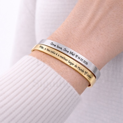 Dois braceletes metálicos, um prateado e outro dourado, com mensagens gravadas no pulso de uma pessoa.
