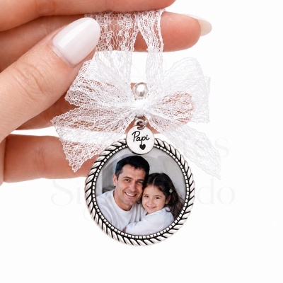 Medalhão redondo prateado com fotografia, laço de renda branco e pendente com texto Papi.