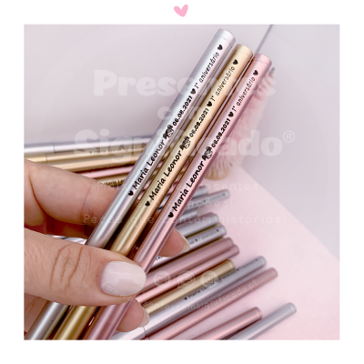 Canetas metálicas prateada, dourada e rosa com texto personalizado, seguradas por mão feminina
