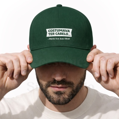 Boné verde escuro com texto branco COSTUMAVA TER CABELO... ...depois tive duas filhas! usado por homem de barba.